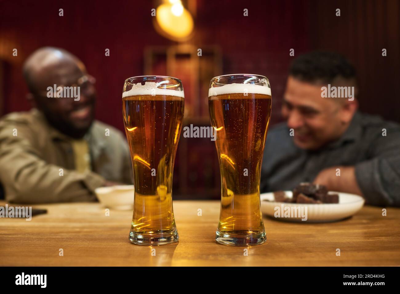 Zwei große Gläser frisches, kühles, schäumendes Bier mit winzigen Blasen im Inneren stehen auf einer Holztheke mit zwei interkulturellen Freunden Stockfoto