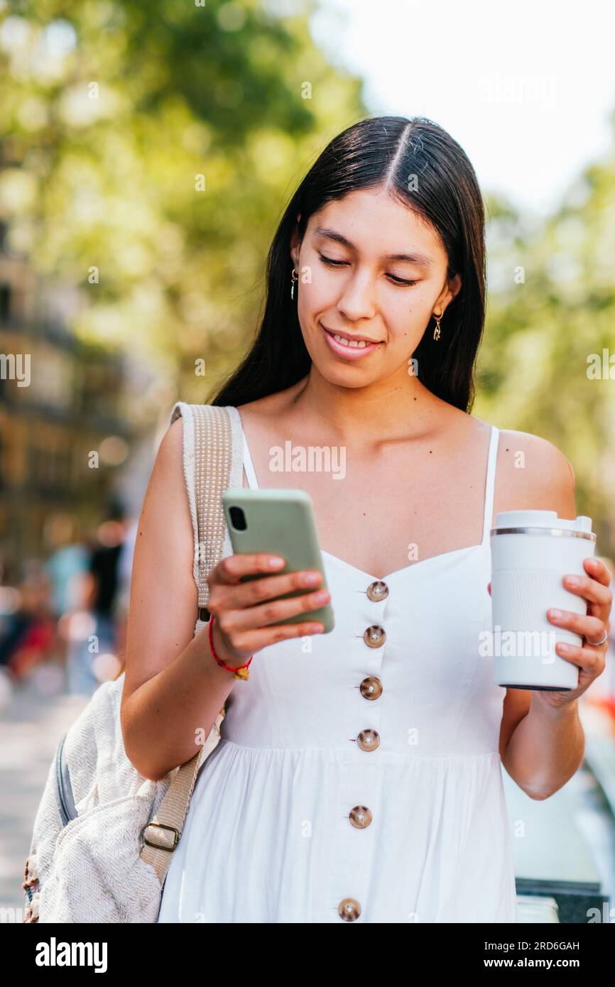 Eine lateinamerikanische Frau in weißem Kleid mit Rucksack und einer Tasse Kaffee, die auf ihrem Smartphone lächelt und auf dem unscharfen Hintergrund des Parks in Barcelona, Spanien, in sozialen Medien surft Stockfoto