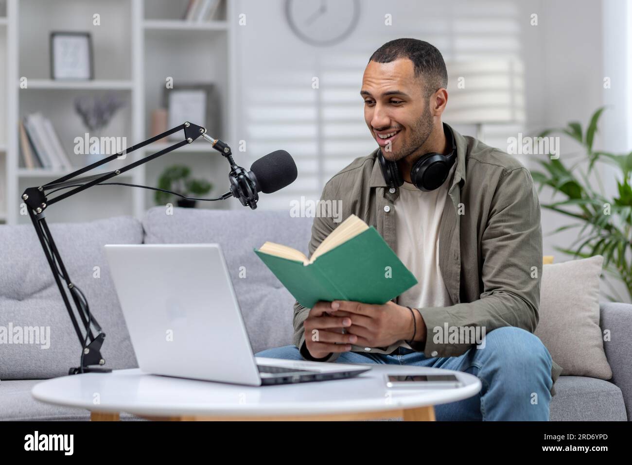 Junger hispanischer Mann mit Kopfhörern, sitzt zu Hause vor einem Mikrofon und Laptop und liest online ein Buch. Stockfoto