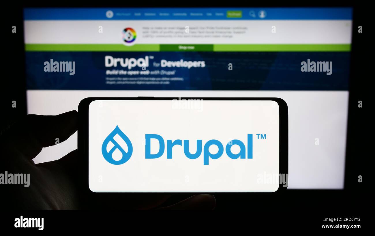 Person, die ein Mobiltelefon mit dem Logo des Content-Management-Systems Drupal auf dem Bildschirm vor der Webseite hält. Konzentrieren Sie sich auf das Display des Telefons. Stockfoto