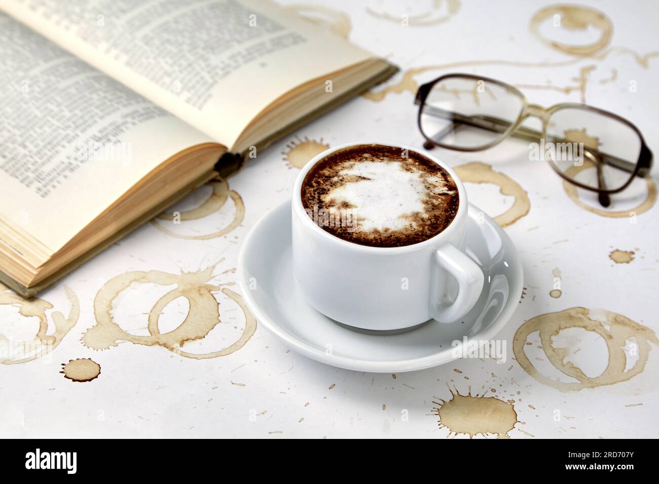 Eine Tasse Cappuccino-Kaffee, Lesegläser und ein Buch auf einem weißen Tisch mit Spuren von Kaffeetassen. Schlaflose Nacht und intensive wissenschaftliche Arbeit vor Exa Stockfoto