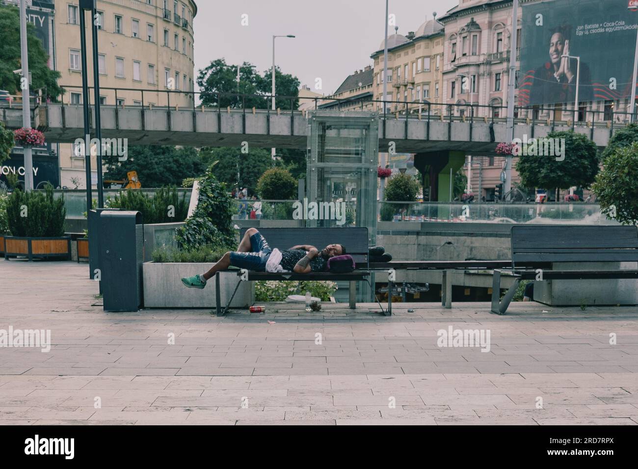 Ein Obdachloser, der auf einer Bank im Stadtzentrum schläft. In Ungarn, insbesondere in Budapest, herrscht anhaltende Besorgnis über Obdachlosigkeit. Aufgrund der hohen Inflation ist die Zahl der Menschen, die auf den Straßen leben, in der Stadt gestiegen. Faktoren wie Armut, Arbeitslosigkeit, psychische Gesundheit und Mangel an bezahlbarem Wohnraum tragen zu dem Problem bei. Die ungarische Regierung und die lokalen Behörden haben verschiedene Maßnahmen zur Bekämpfung der Obdachlosigkeit ergriffen. Dazu gehören die Bereitstellung von Notunterkünften, soziale Dienste und Programme zur Wiedereingliederung in die Gesellschaft. Nicht-staatliche Stockfoto