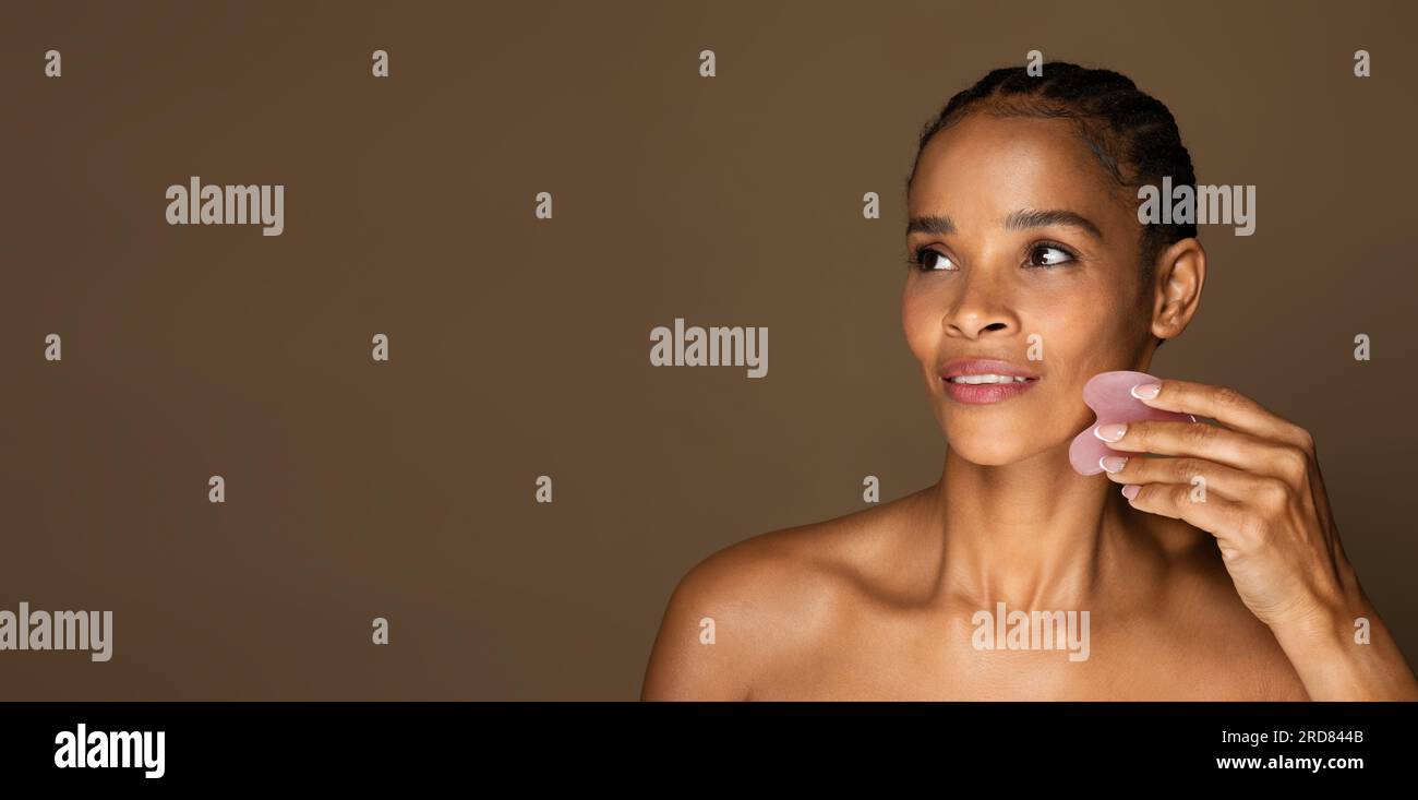 Wunderschöne schwarze Frau mittleren Alters, die eine Gesichtsmassage mit Gua-Sha-Steinkratzer macht, Panorama mit freiem Platz, Banner Stockfoto