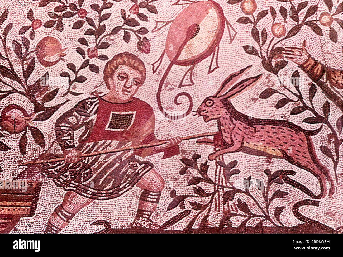 Dieses Foto eines Mosaiks mit einer Jagdszene (Szene eines jungen Mannes/Jungen, der einen Hasen jagt) wurde im Sommer 1970 auf der Piazza Armerina in Sizilien aufgenommen. Die Piazza Armerina beherbergt die römische Villa del Casale und ihre berühmten Mosaiken, die „besten Mosaike in situ in der römischen Welt“, wie von der UNESCO beschrieben, die sie 1997 in ihre Liste des Weltkulturerbes aufgenommen hat. Villa Romana war eine üppige Patrizier-Residenz, die im Zentrum eines riesigen Latifundiums (Landwirtschaftsgelände) am Ende des 4. Jahrhunderts n. Chr. erbaut wurde. Es wird angenommen, dass es einem Mitglied der römischen Senatorenaristokratie gehörte, die Handel treibt Stockfoto