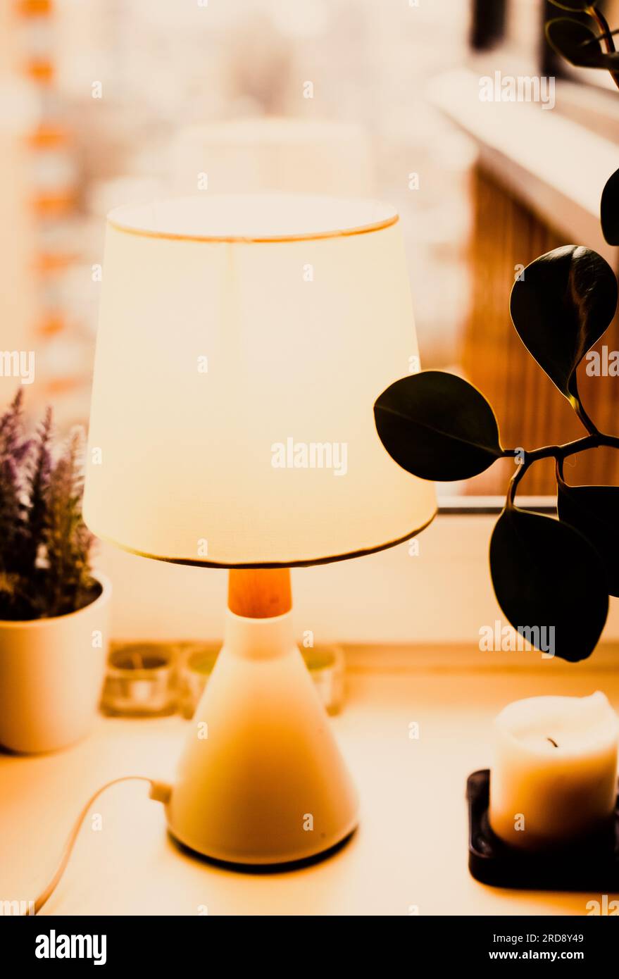 Eine gemütliche Innenatmosphäre mit einer Schreibtischlampe, Zimmerpflanzen und einer Kerze, die auf einer Fensterbank ruht. Wärme, Ruhe und ein einladendes Ambiente. Stockfoto Eine gemütliche Innenatmosphäre mit einer Schreibtischlampe, Zimmerpflanzen und einer Kerze, die auf einer Fensterbank ruht. Wärme, Ruhe und ein einladendes Ambiente. Stockfoto