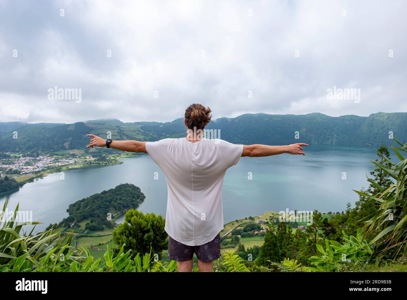 Junger Mann in weißem T-Shirt mit ausgestreckten Armen und Blick auf den See von Seven Cities Lake „Lagoa das Sete Cidades“. Die Insel Sao Miguel auf den Azoren. Stockfoto