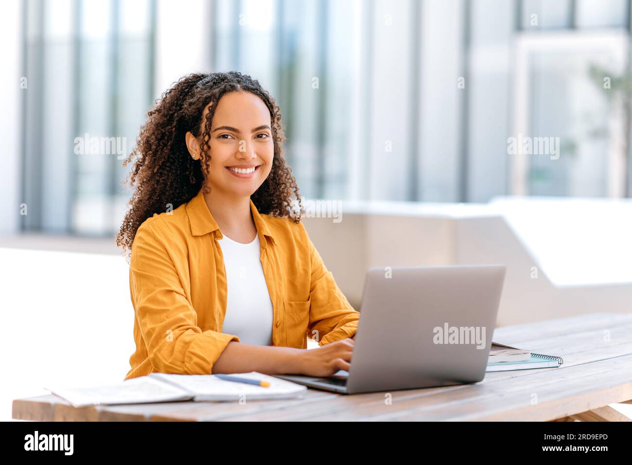 Foto eines erfolgreichen, lockigen Mädchens, brasilianischer oder lateinamerikanischer Nationalität, Auslagerung von Mitarbeitern oder Studenten, Verwendung eines Laptops zum Arbeiten oder Lernen im Internet, während er draußen sitzt, in die Kamera schaut, freundlich lächelt Stockfoto