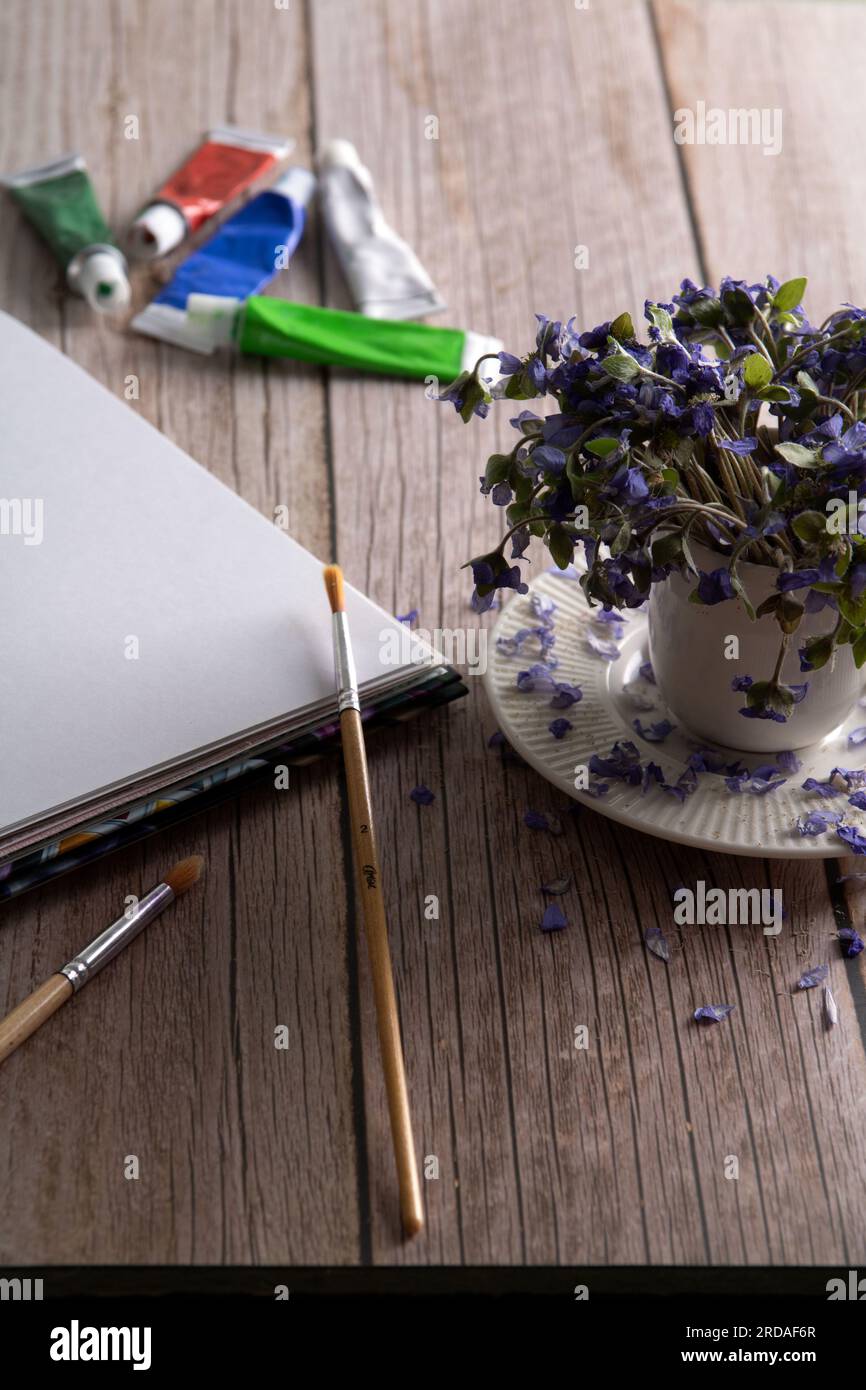 Foto einer getrockneten Hepatica, die in einem Becher neben einem weißen Blatt Papier, Farben und Pinseln auf dem Tisch steht. Stockfoto