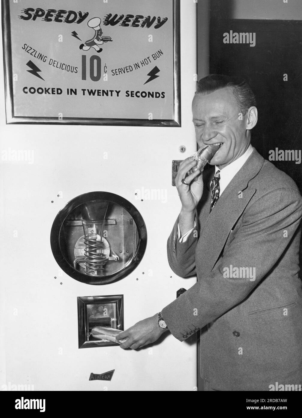HIER KOMMT DER ELEKTRONISCHE HOT DOG New York... als wäre das Leben nicht kompliziert genug, kommt das Gerät zum Dispensieren des elektronisch gekochten Frankfurter. Es heißt The Radio Chef was was was was wurde heute in New york vorgeführt, mit Funnyman Sammy Kaye als einem der ersten Kunden. Kaye wird hier gezeigt, wie er einen Rotschopf abbricht, während ein anderer bereit zur Zerstörung auftaucht. Die Frankfurter sind in Zellophan eingewickelt und in eine Ofenfrische Rolle eingehüllt, bereit für den Verbraucher, der einen Cent in den unvermeidlichen Slot einzahlt. Schnell, winzig. Köstliche 10 Cent in einem heißen Brötchen. In zwanzig sekunden zubereitet Stockfoto