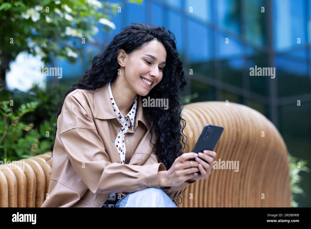 Fröhliche, schöne Frau, die vor dem Bürogebäude auf einer Bank sitzt, erfolgreiche lateinamerikanische Geschäftsfrau, die telefoniert, Nachrichten im Internet liest und online surft, ihr Smartphone in der Hand hält. Stockfoto