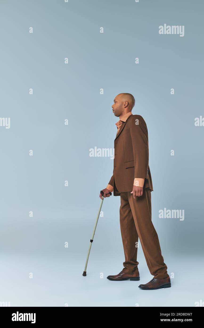 Echte Menschen, voller Blick auf einen afroamerikanischen Mann mit Myasthenia gravis, der sich auf einem Gehstock auf grauem Hintergrund lehnt, dunkle gehäutete Person im Anzug, Stockfoto