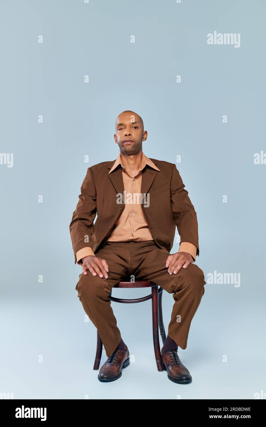 Echte Menschen, voller Länge, ein kühner afroamerikanischer Mann mit Myasthenia gravis, der auf einem Holzstuhl auf grauem Hintergrund sitzt, dunkle, gehäutete Person im Anzug Stockfoto