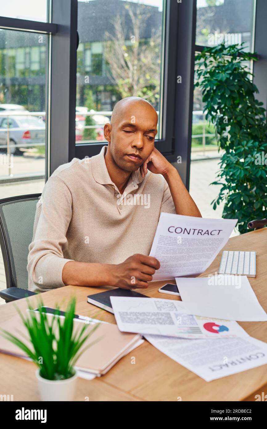 Inklusion und Vielfalt, afroamerikanischer Mann mit Myasthenie Krankheit, der auf Vertrag schaut, dunkel gehäuteter Büroangestellter in lässiger Kleidung am Schreibtisch sitzen Stockfoto