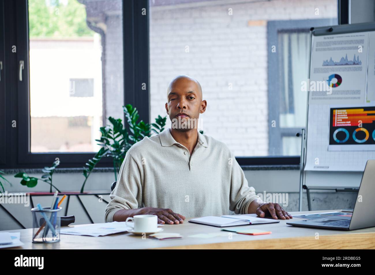 Chronische Krankheit, kühner afroamerikanischer Geschäftsmann mit Augensyndrom, der in der Nähe des Laptops auf dem Schreibtisch auf die Kamera schaut, Myasthenia gravis, dunkelhäutiges Büro Stockfoto