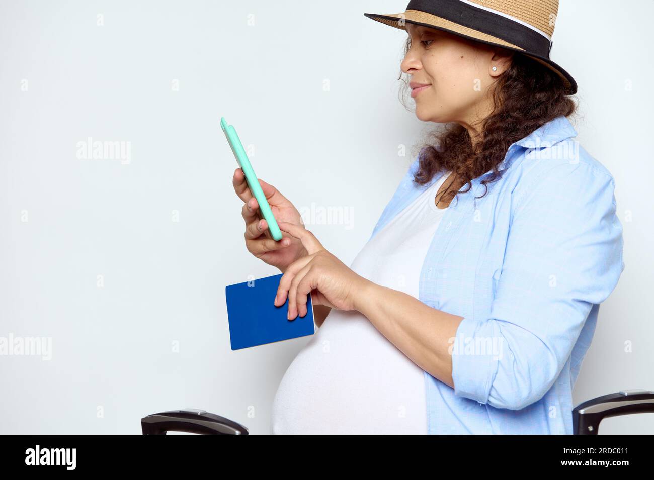 Seitliches Porträt einer lockigen, multiethnischen, schwangeren Frau mit Strohhut, die mobile Anwendungen auf dem Smartphone überprüft, ein Passdokum in der Hand hat Stockfoto