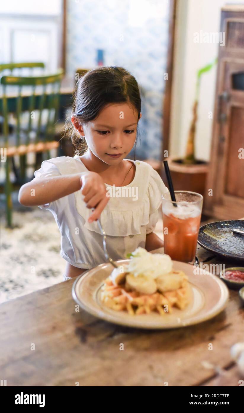 Kleines asiatisches Mädchen, das süße belgische Waffeln mit Banane und Sahne in einem Café isst Stockfoto