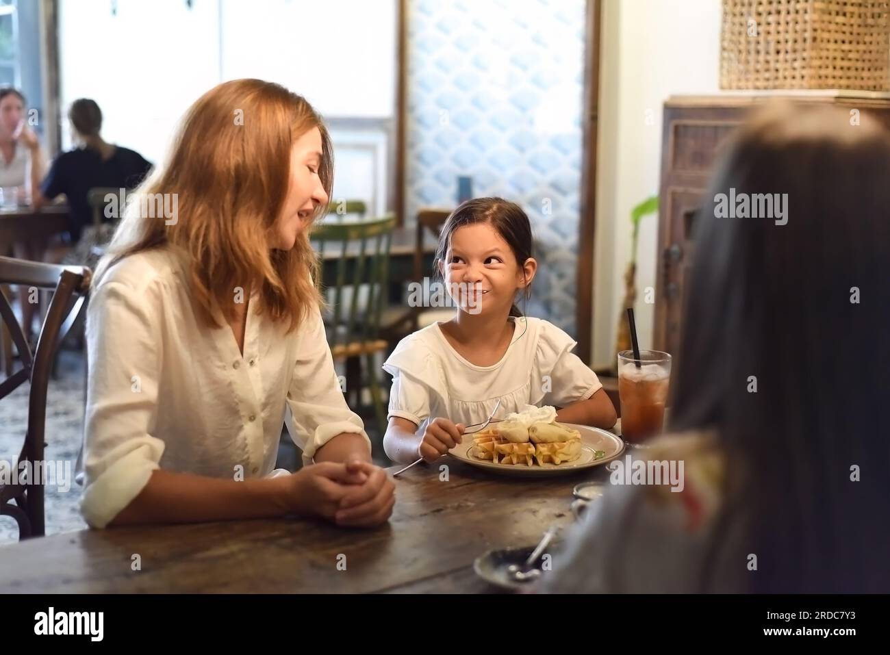 Kleines Mädchen und ihre Mutter essen süße belgische Waffeln mit Banane und Sahne in einem Café, Tochter macht ein lustiges Gesicht Stockfoto