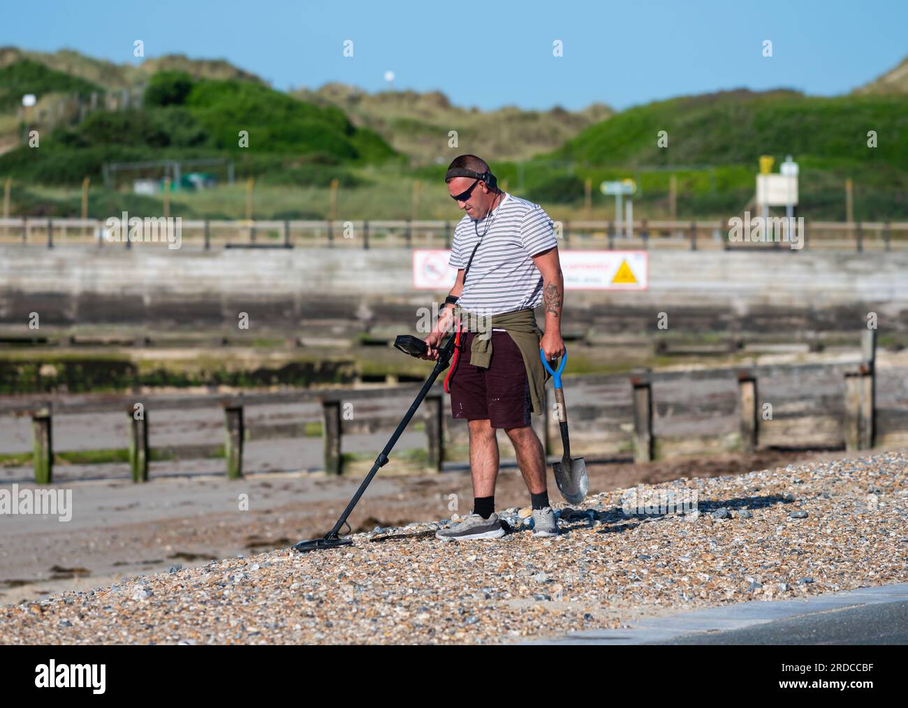 Mann am Kieselstrand mit Metalldetektor, der nach verlorenen oder wertvollen Gegenständen sucht, am Meer in Großbritannien. Metalldetektorin. Stockfoto