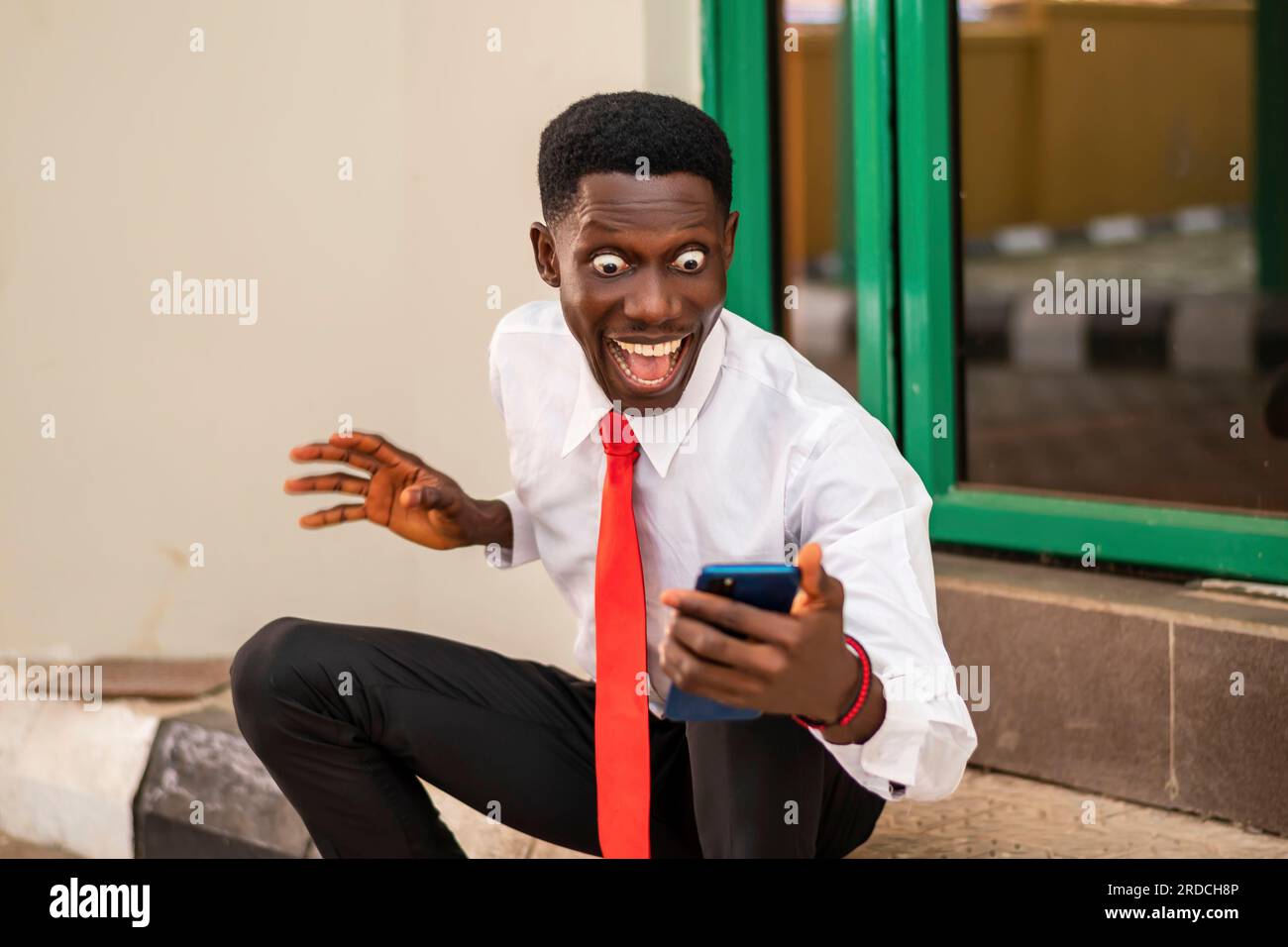 Foto eines jungen Afrikaners, der aufgeregt aussah, während er ein Handy in einem weißen Hemd und einer roten Krawatte hielt Stockfoto