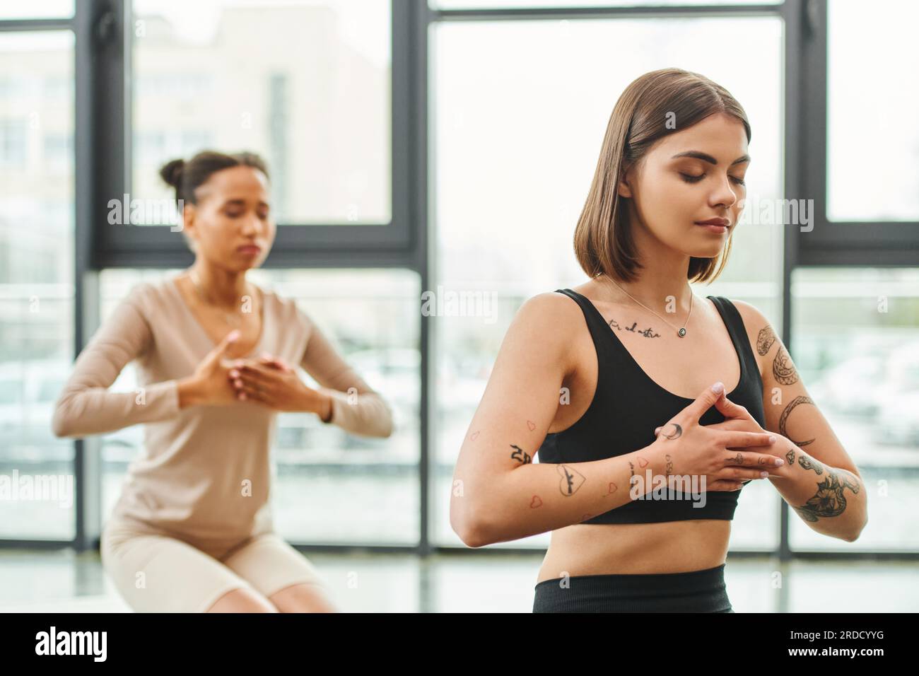 Junge und tätowierte Frau meditiert mit geschlossenen Augen und Händen auf der Brust während des Yoga-Kurses in der Nähe einer afroamerikanischen Freundin auf verschwommenem Hintergrund, inne Stockfoto