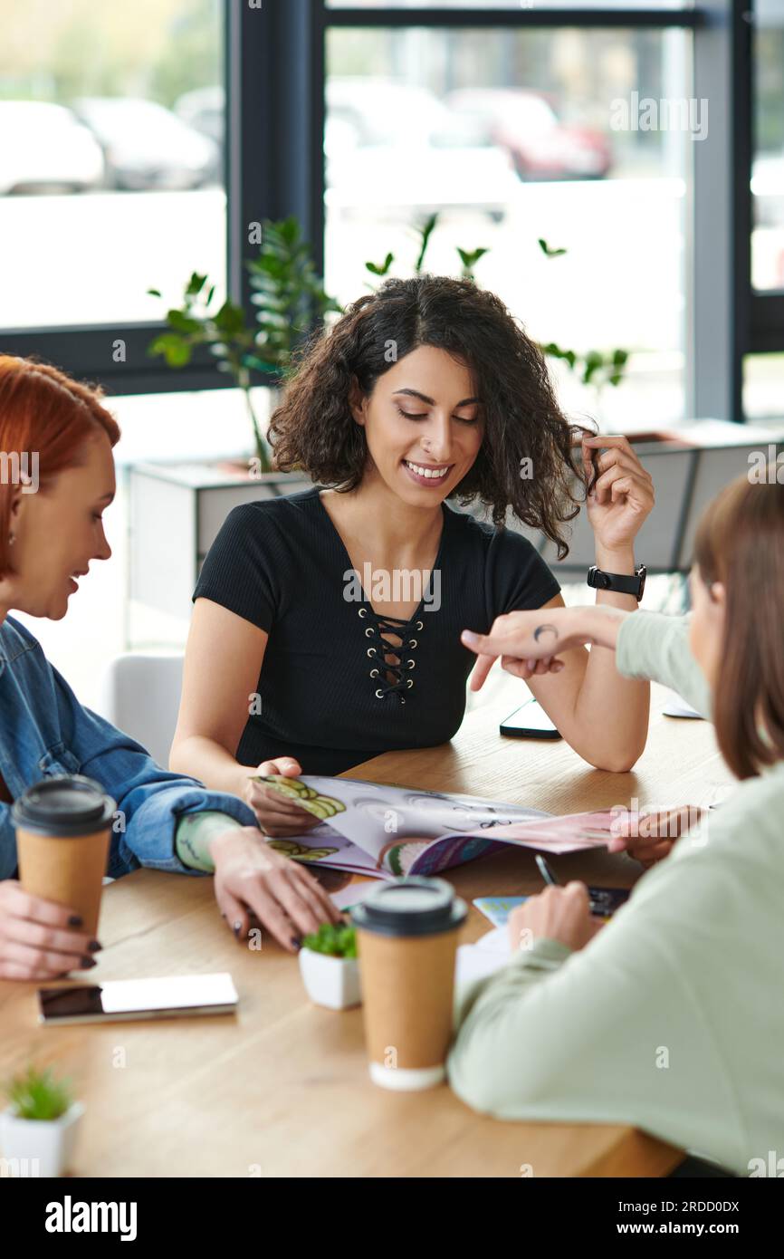 Junge Frau, die auf ein Magazin zeigt, lächelt multikulturelle Freundinnen und Pappbecher mit Kaffee, die man im Frauenclub auf den Tisch bekommt, gemeinsame Interessen und Stockfoto