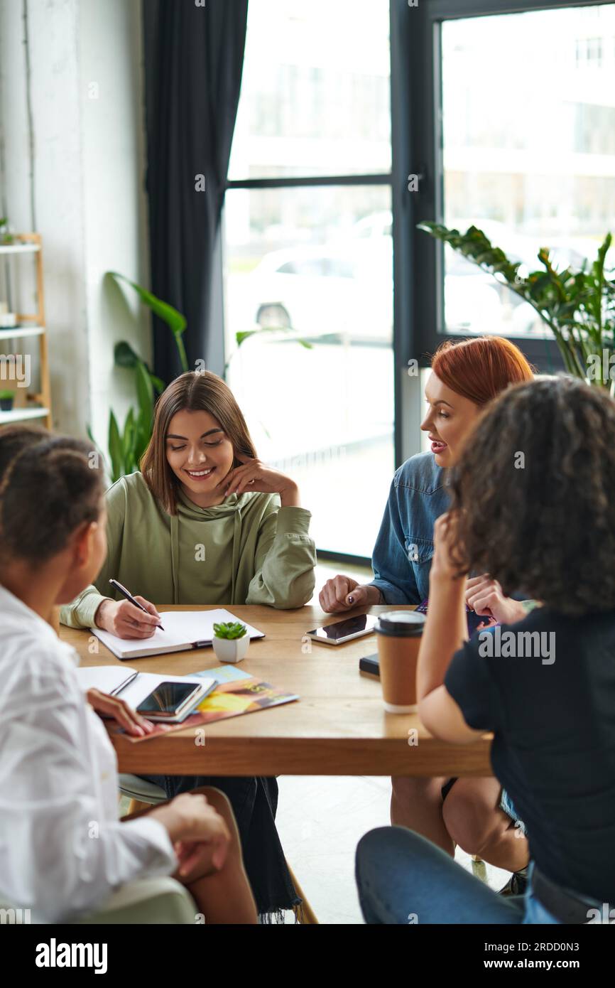 Ein zufriedenes Mitglied des Interessenverbands der Frauen schreibt in Notizbücher in der Nähe von Freundinnen, Mobiltelefonen, Kaffee zum Mitnehmen und Zeitschriften auf dem Tisch, Wissensaustausch A Stockfoto