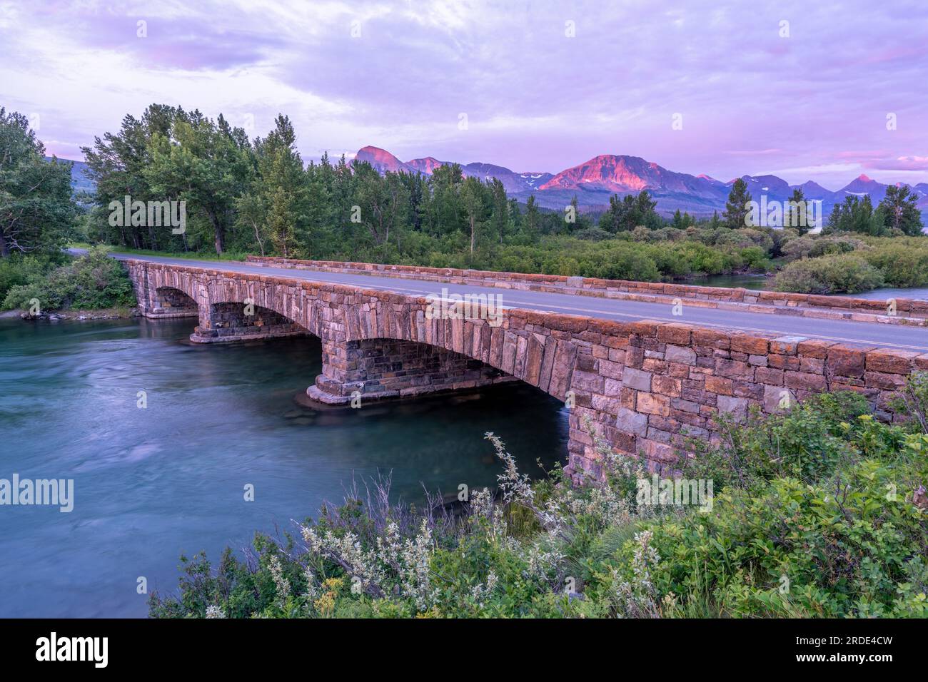 Eine Steinbrücke, die einen breiten Fluss mit Bäumen im Hintergrund im sanften Licht vor Sonnenaufgang überspannt, Saint Mary Lake Bridge, Glacier National Stockfoto