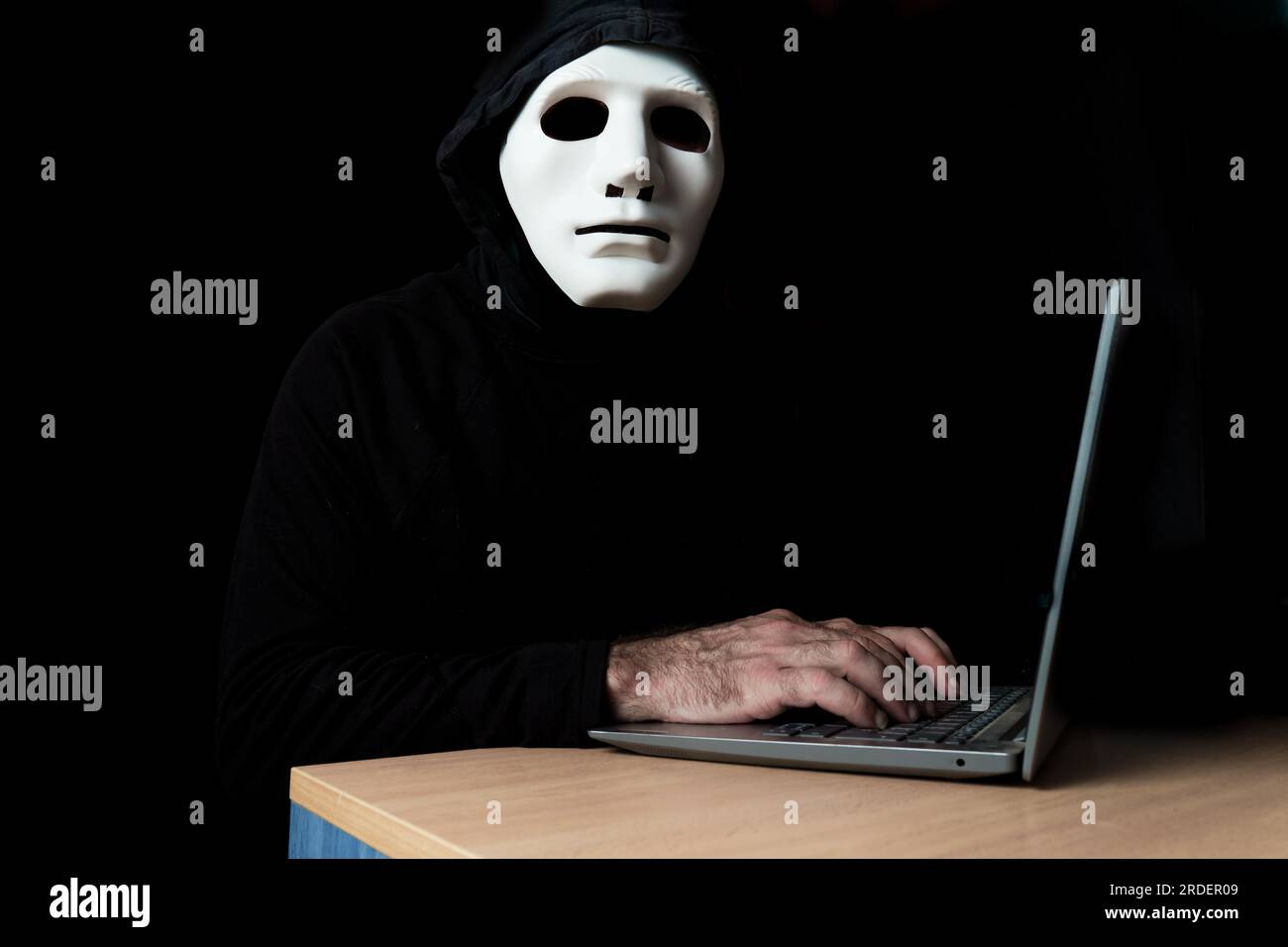 Hacker in Schwarz gekleidet mit Maske und Kapuze auf einem Computer, während er sich das Cyber-Crime-Konzept der Kamera ansieht Stockfoto