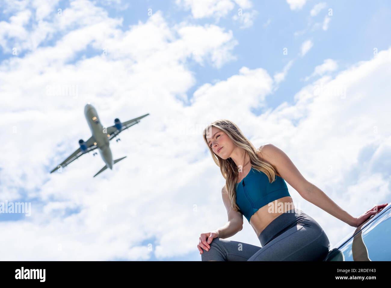 Ein schönes blondes Modell genießt einen Sommertag, während ein kommerzielles Flugzeug über dem Himmel fliegt Stockfoto