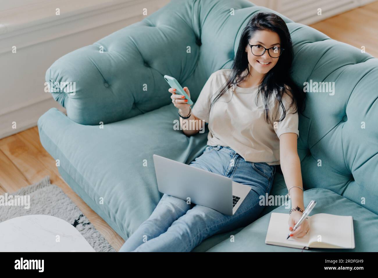 Fröhliche Freiberuflerin in lässiger Kleidung, schreibt in Notizblock, benutzt Laptop, auf dem Sofa. Der Schüler bereitet die Projektarbeit zu Hause vor. Stockfoto