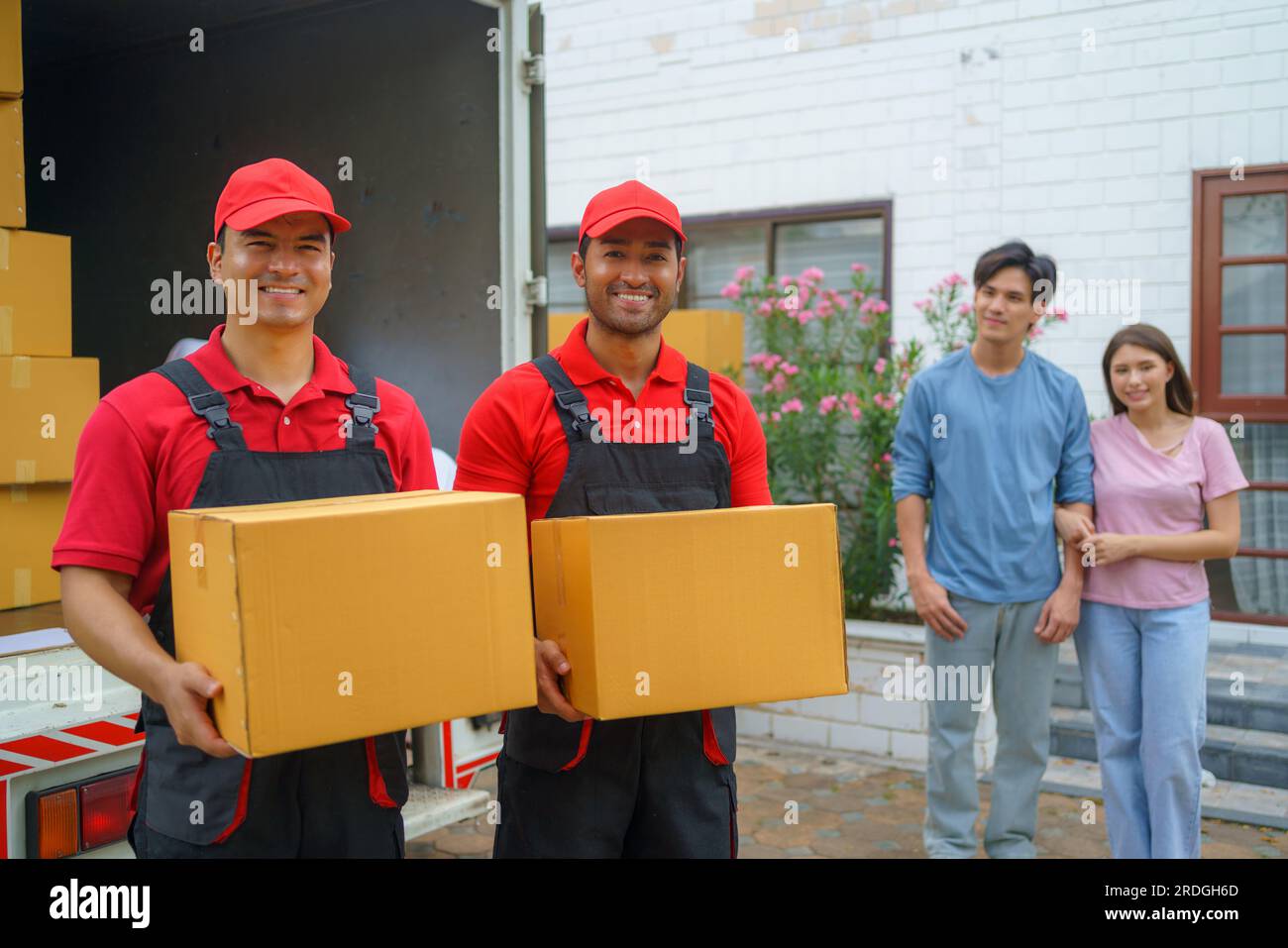 Ein asiatisches Paar steht zusammen und beobachtet mit Vorfreude, wie Arbeiter in ihr neues Zuhause ziehen. Spannung füllt die Luft, wie sie eifrig erwarten Stockfoto