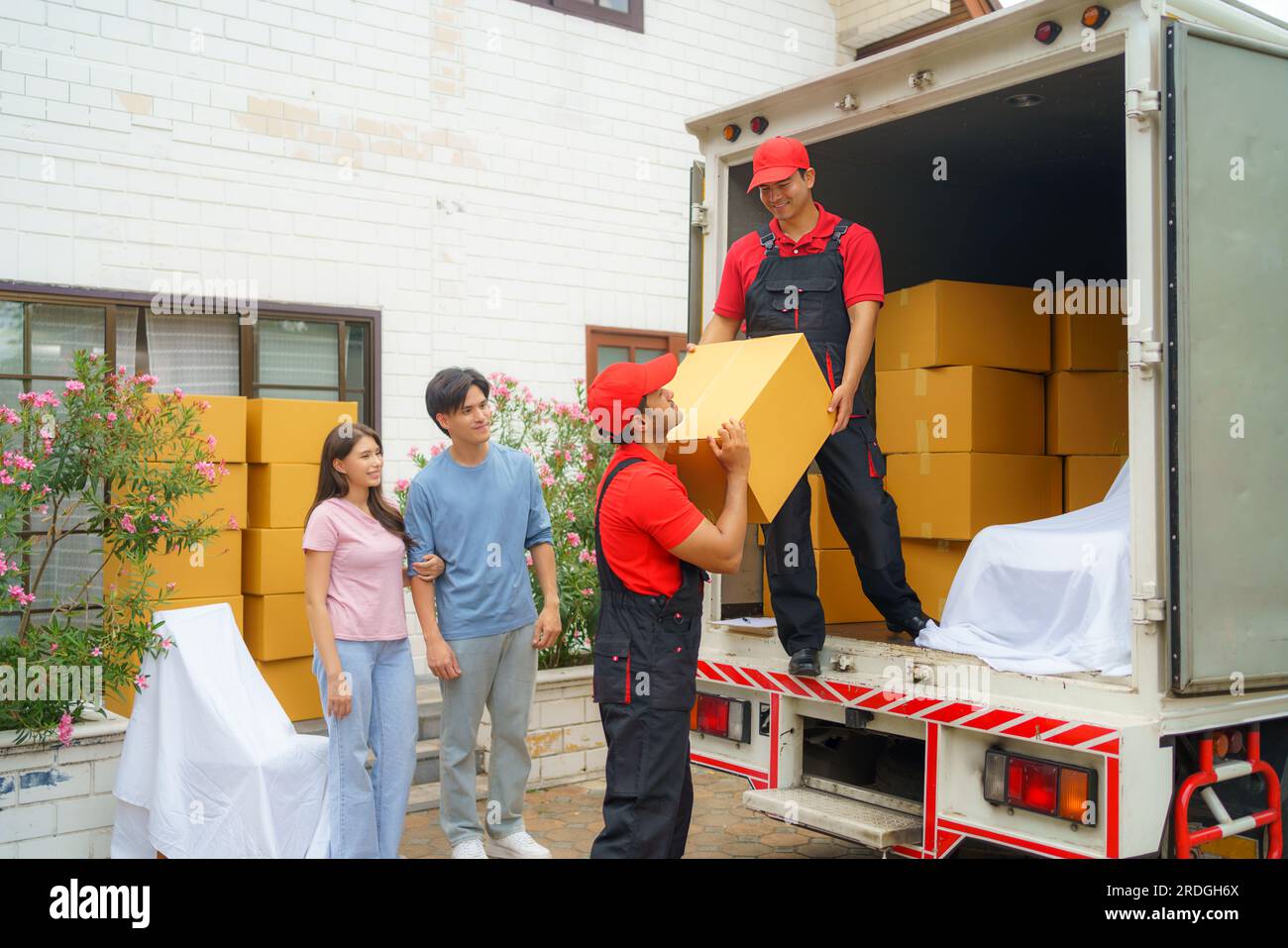Ein asiatisches Paar steht zusammen und beobachtet mit Vorfreude, wie Arbeiter in ihr neues Zuhause ziehen. Spannung füllt die Luft, wie sie eifrig erwarten Stockfoto