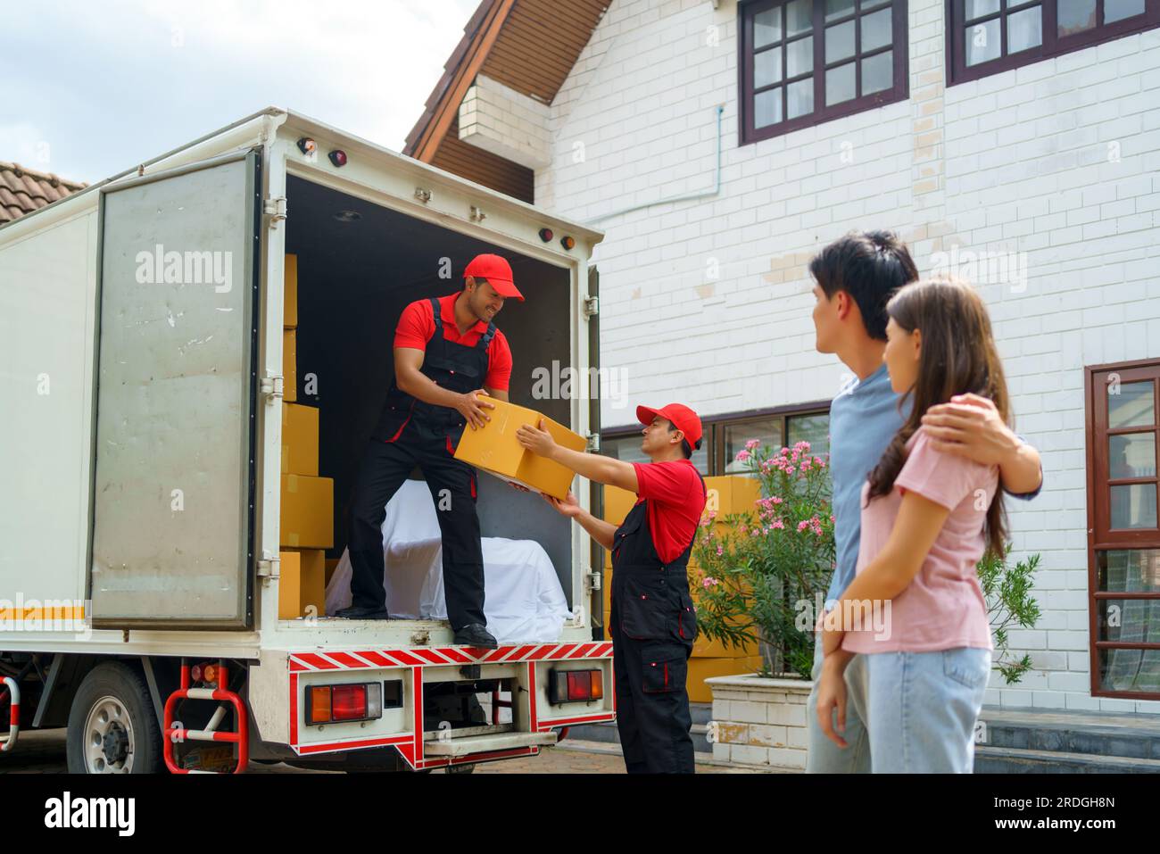 Ein asiatisches Paar steht zusammen und beobachtet mit Vorfreude, wie Arbeiter in ihr neues Zuhause ziehen. Spannung füllt die Luft, wie sie eifrig erwarten Stockfoto
