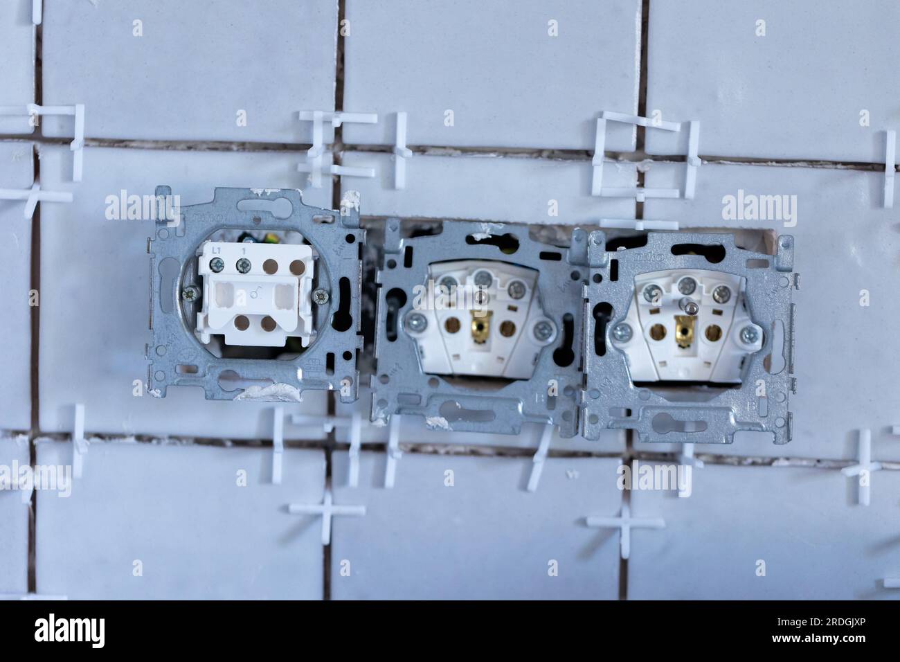 Ein Porträt mit einem Lichtschalter und zwei elektrischen Wandsteckdosen ohne Schutzkappen in einer neu gefliesten Wand, immer noch mit den weißen Kunststofffugen Stockfoto