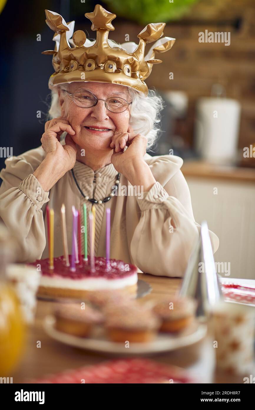 Eine geliebte Oma sitzt am Tisch, ihr Gesicht schmückt mit einem strahlenden Lächeln, das ein Leben voller Liebe und Weisheit widerspiegelt. Vor ihr ist Geburtstag Stockfoto