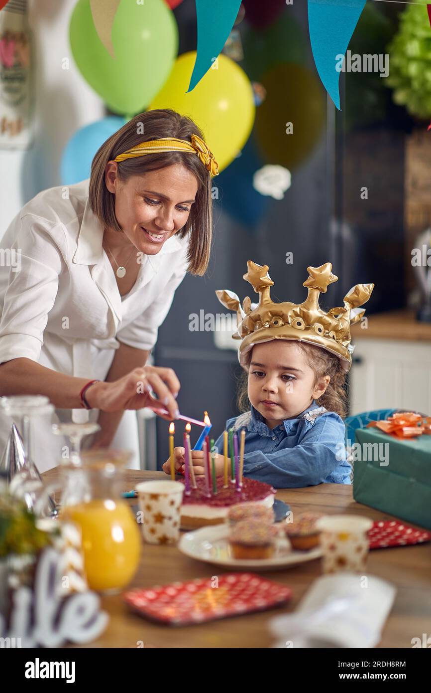 Schönes Geburtstagskind mit goldener Krone, das sich etwas wünscht, während Mutter ihre Kerzen auf dem Kuchen anzündet. Heim, Familie, Festungskonzept. Stockfoto