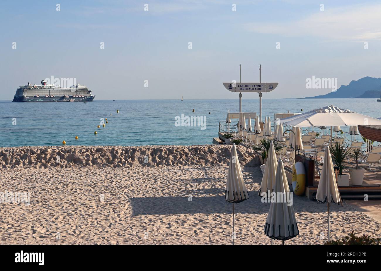 Am frühen Abend fällt die Sonne auf einen Teil des Carlton Beach Club in Cannes, bevor die Saison beginnt. Draußen auf dem Meer liegt ein riesiges TUI-Kreuzfahrtschiff vor Anker. Stockfoto