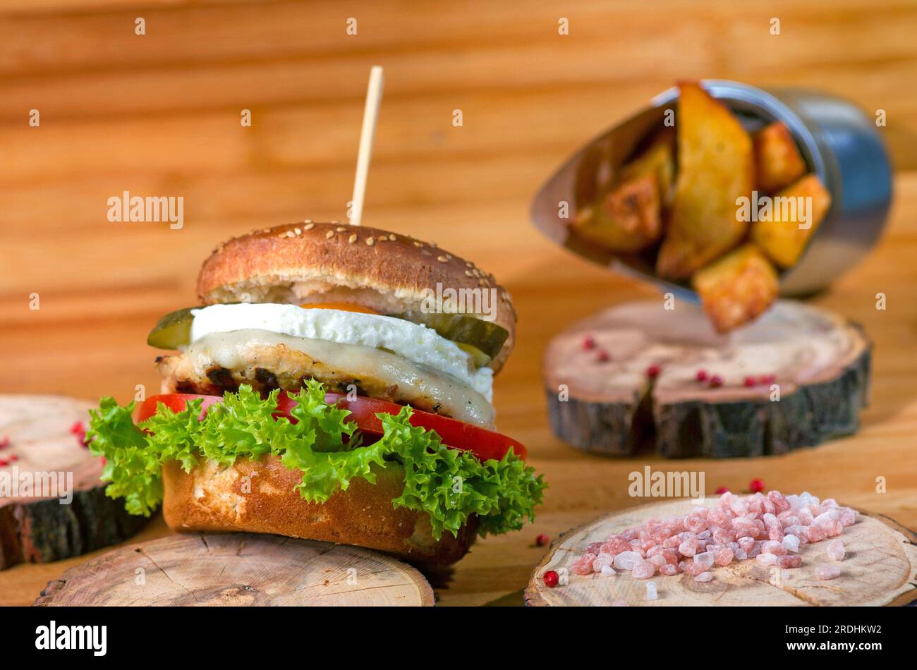 Saftiger Burger mit gesalzenem Fleisch und Ei auf einer Holzplatte mit Salzstücken an der Seite und gebackenen Kartoffeln im Hintergrund Stockfoto