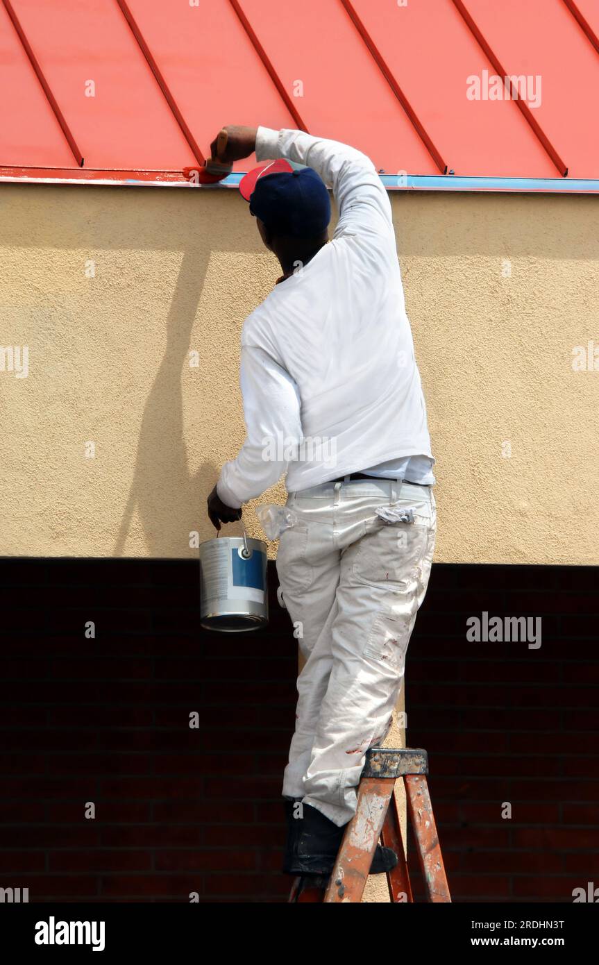 Der Mann steht auf der obersten Stufe der Leiter, während er die Dachlinie am Bürogebäude in der Innenstadt von Albuquerque, New Mexico, lackiert. Er hält eine Farbdose und Sprea in der Hand Stockfoto