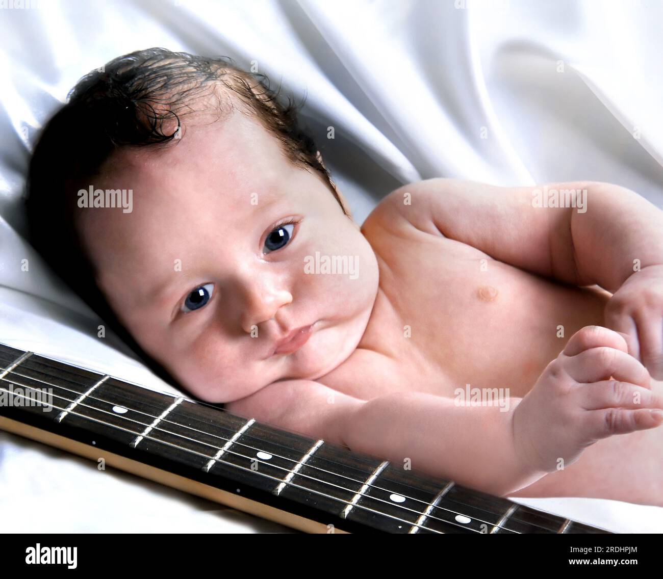 Das Neugeborene liegt neben der Gitarre seines Vaters. Das musikalische Training sollte früh beginnen, und dieser Elternteil möchte die musikalische Langeweile seines Kindes beeinflussen. Stockfoto