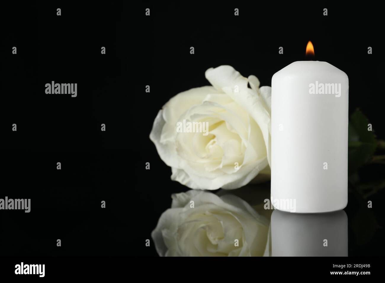 Weiße Rose und brennende Kerze auf schwarzer Spiegeloberfläche bei Dunkelheit, Nahaufnahme mit Platz für Text. Begräbnissymbole Stockfoto