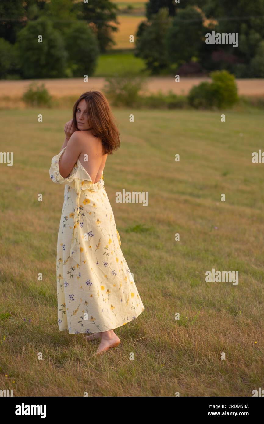 Eine junge, schöne Dame geht barfuß auf einem Feld bei Sonnenuntergang. Ein Mädchen in einem wunderschönen gelben Kleid posiert bei Sonnenuntergang auf einem Feld. Stockfoto