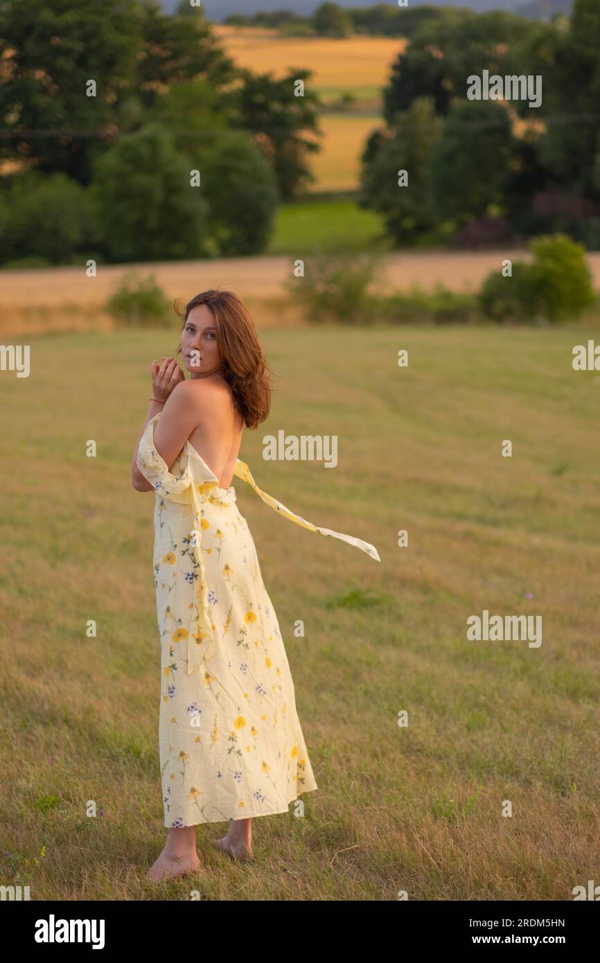 Eine junge, schöne Dame geht barfuß auf einem Feld bei Sonnenuntergang. Ein Mädchen in einem wunderschönen gelben Kleid posiert bei Sonnenuntergang auf einem Feld. Stockfoto