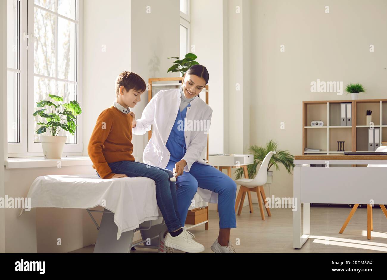 Neurologe, der den kleinen Jungen untersucht, der in der Klinik mit dem Hammer auf das Knie klopft. Stockfoto