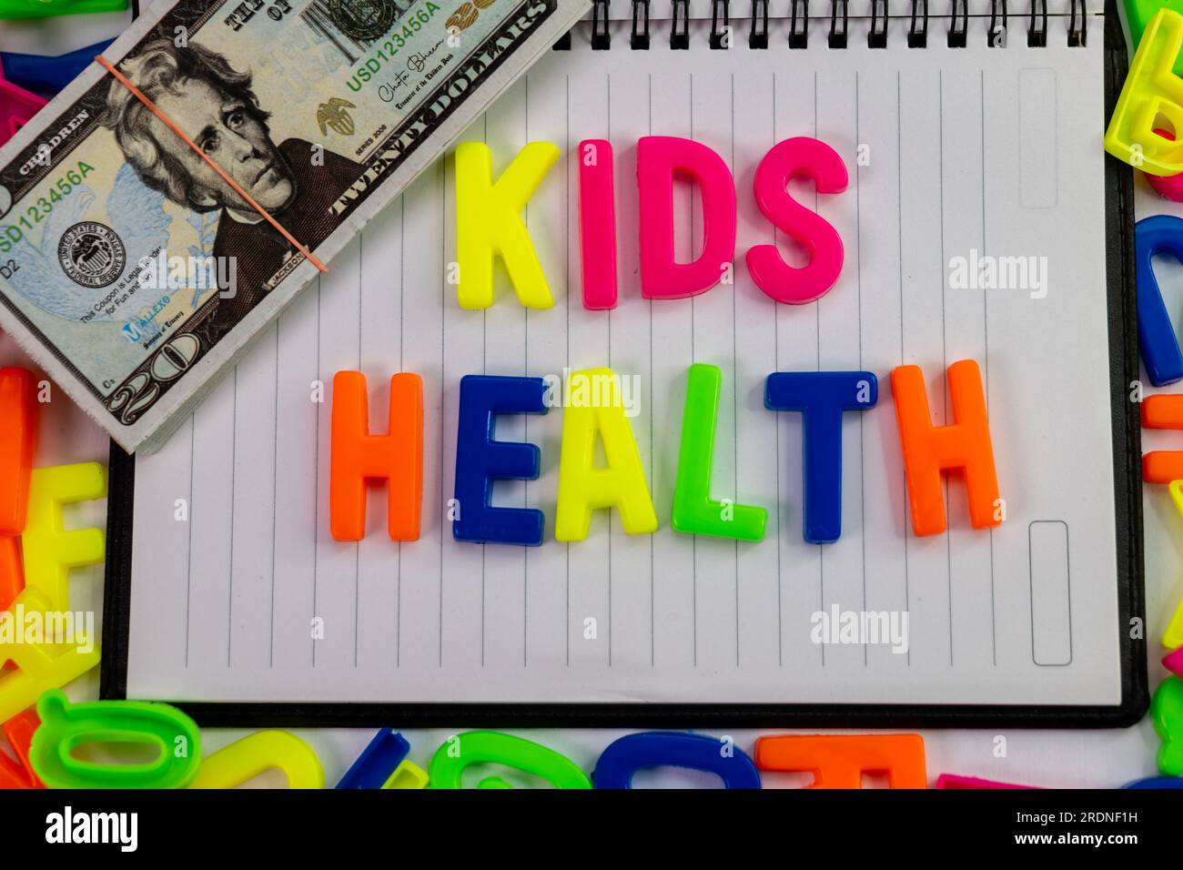 Eine Nahaufnahme von bunten Buchstabenmagneten, die die Gesundheit von Kindern auf einem einfachen Stück Milchprodukte erklären, mit einem Stapel Bargeld oben drauf. Stockfoto