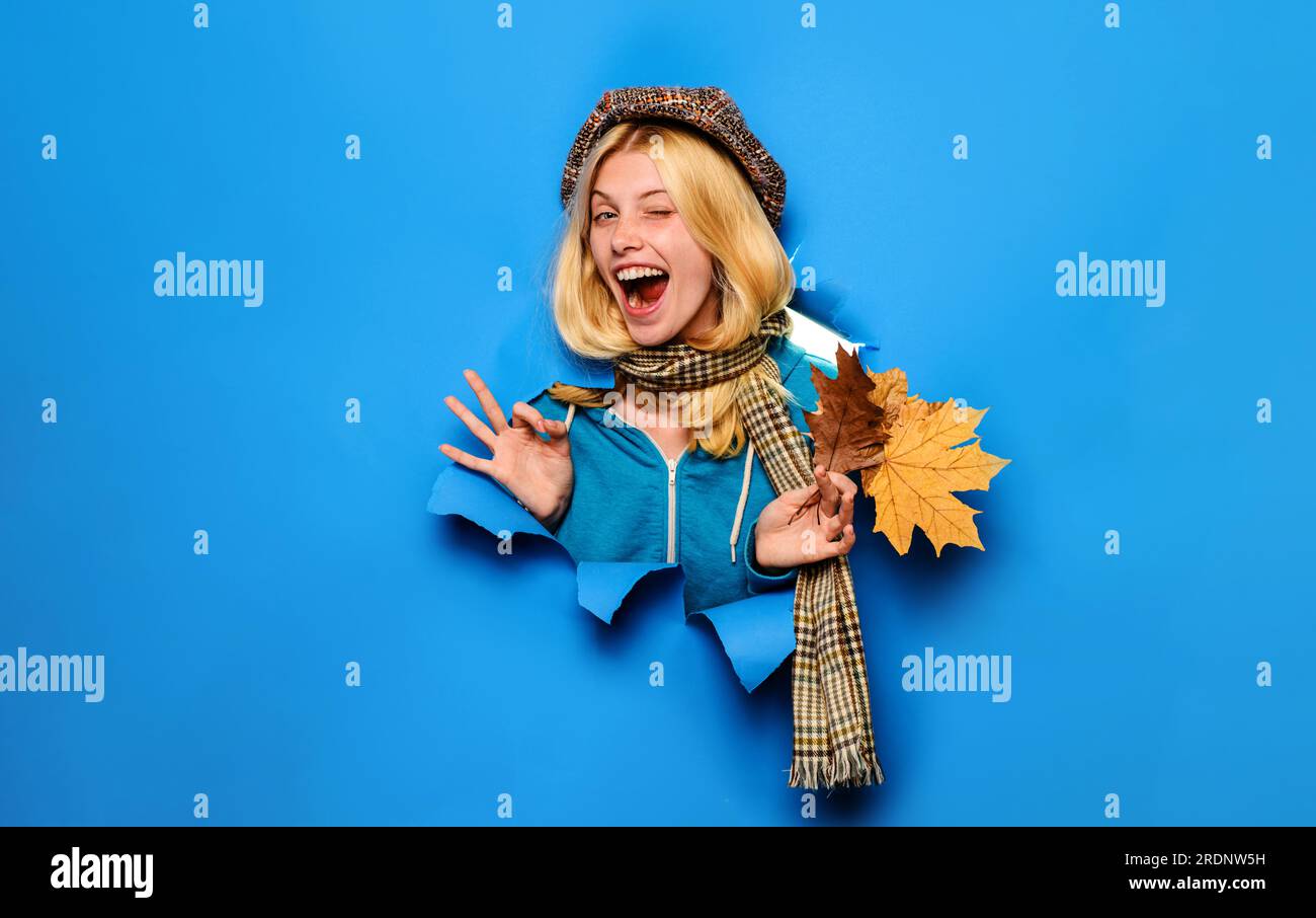 Blinkendes Herbstmädchen mit Mütze und Schal, das ein „OK“-Schild zeigt. Glückliche schöne Frau mit Herbstblättern, die durch das Papierloch blickt. Herbstverkäufe. Rabatt Stockfoto