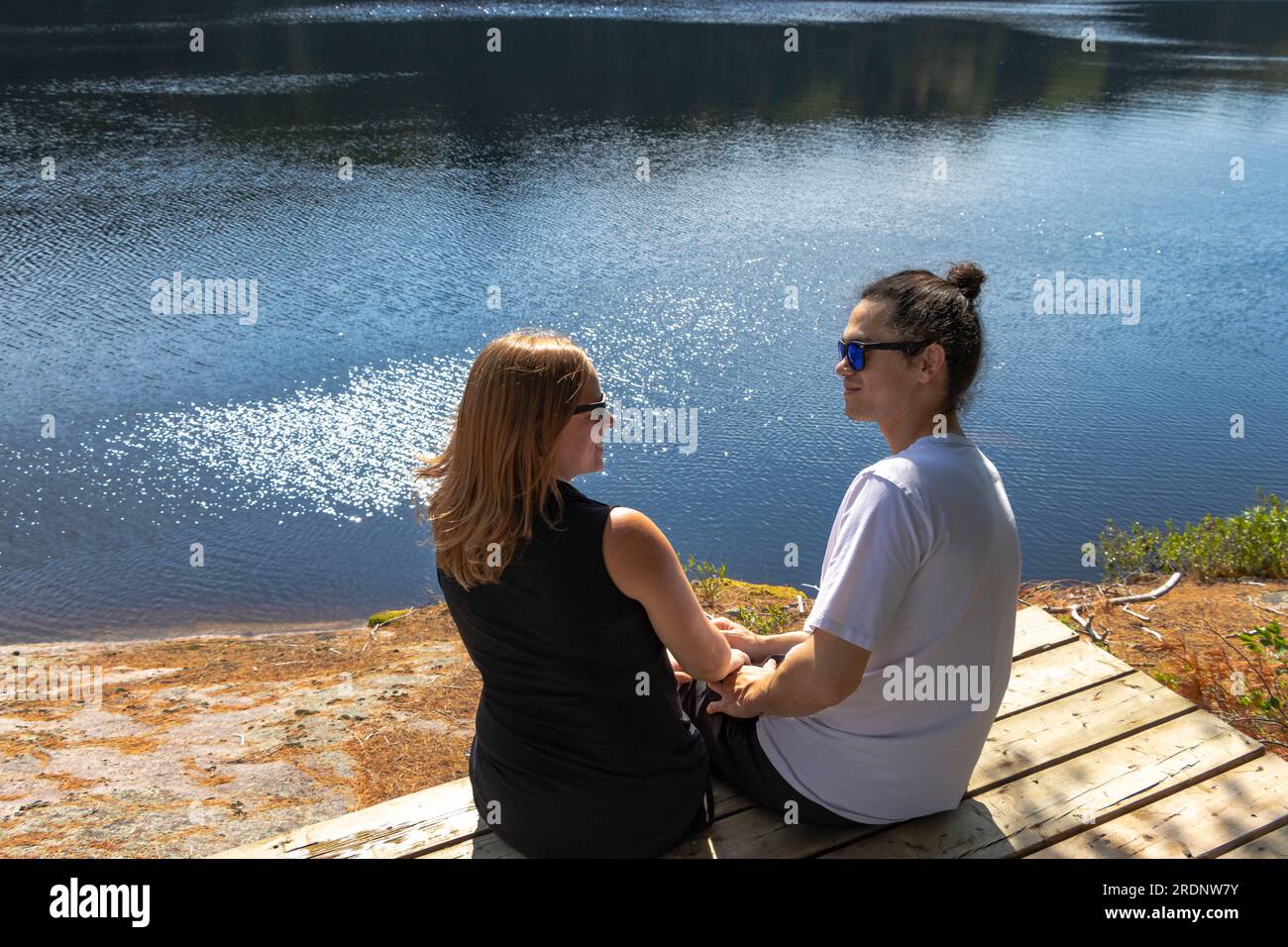Gemischtes Paar, das auf dem Dock am Wasser im Muskoka-Wald in der Natur sitzt Stockfoto