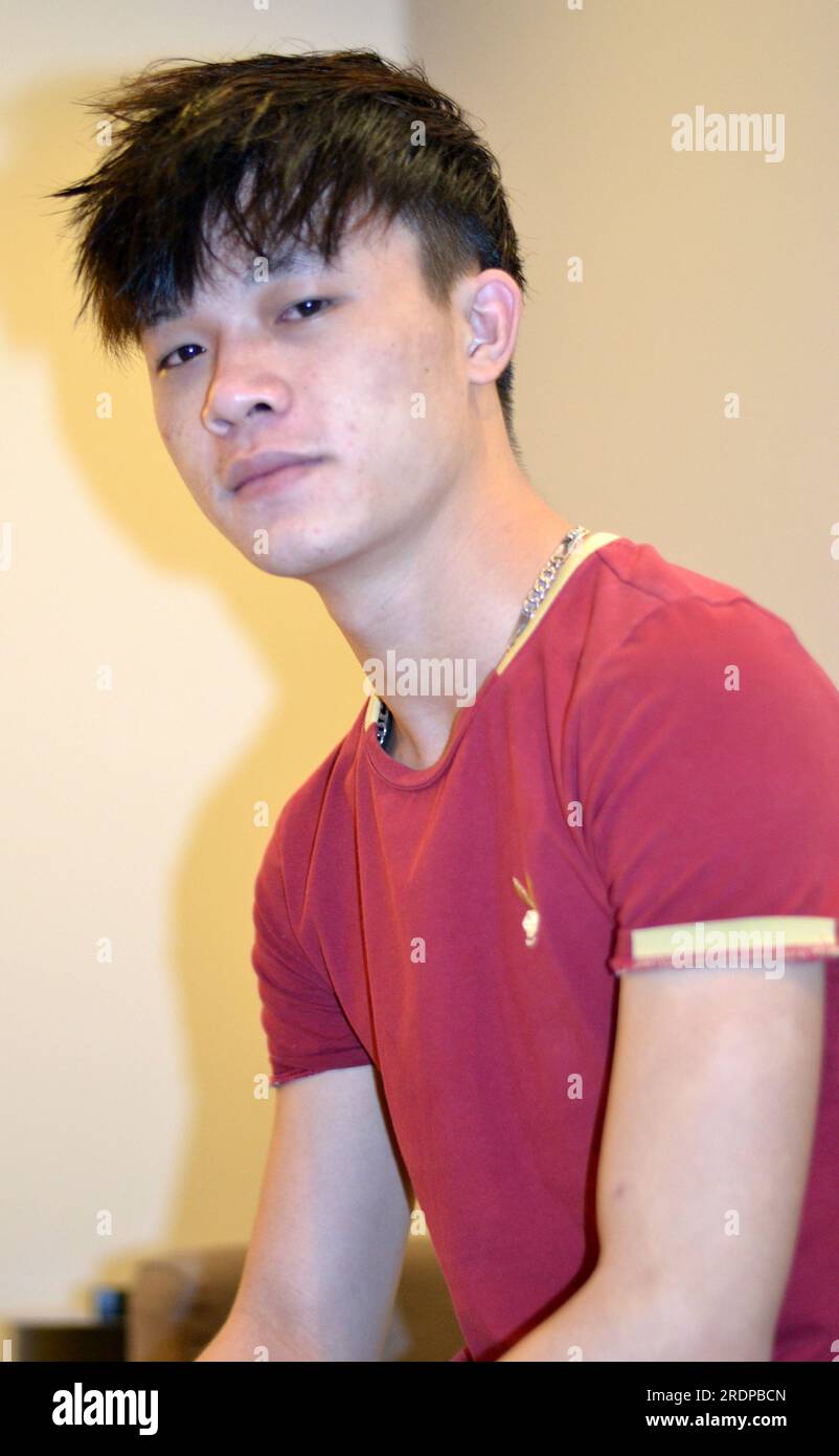 Porträt eines gutaussehenden, jungen, vietnamesischen, asiatischen Mannes, der ein rotes ärmelloses Hemd trägt und in die Kamera schaut, dunkle Haare, Fransen, etwa 20 Jahre alt, Kopierbereich auf hellem Hintergrund, Schatten an der Wand vom verwendeten Blitz Stockfoto