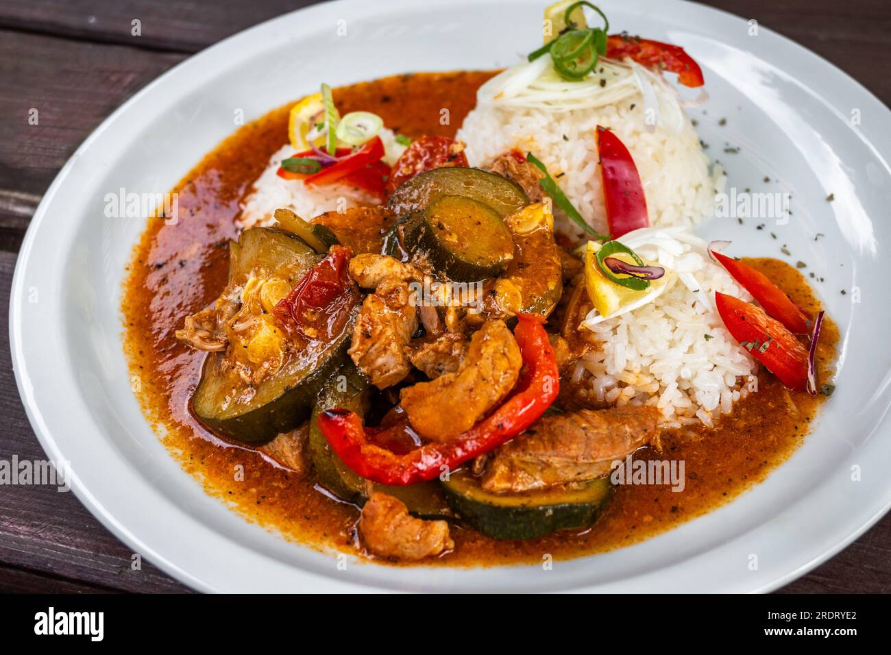 Schweinefleisch mit Gemüse (Zucchini, Zwiebeln, Knoblauch und roter Pfeffer) in scharfer roter Sauce mit Reis auf weißem Teller, Nahaufnahme. Stockfoto