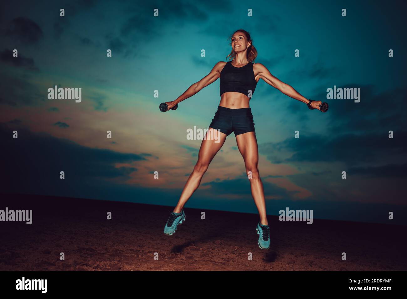 Junge Fitness-Frau, die mit Kurzhanteln im Hintergrund des Dämmernachlichthimmels springt Stockfoto